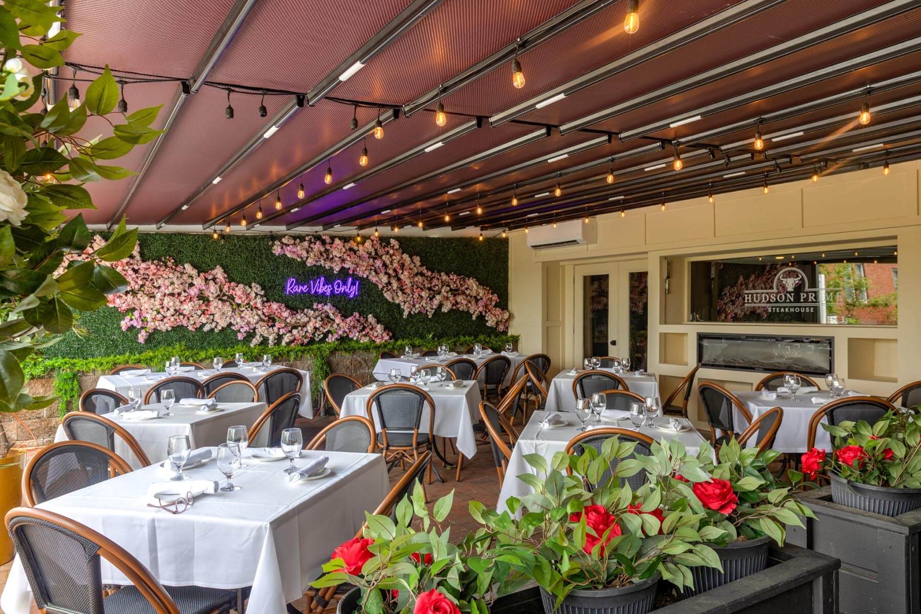 Elegant covered outdoor dining area with white tablecloths, wooden chairs, hanging string lights, and a flower wall with neon sign reading 'Rare Vibes Only'.